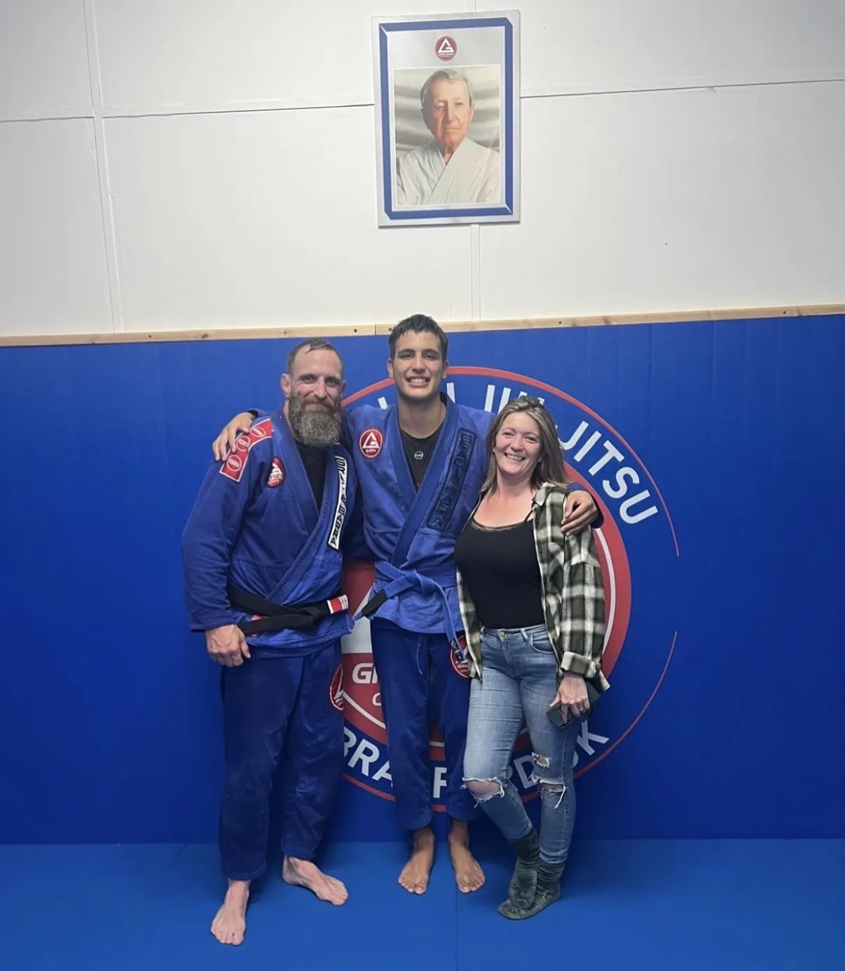 A young athlete at a belt promotion on the mats at the academy, standing between a coach and a family member