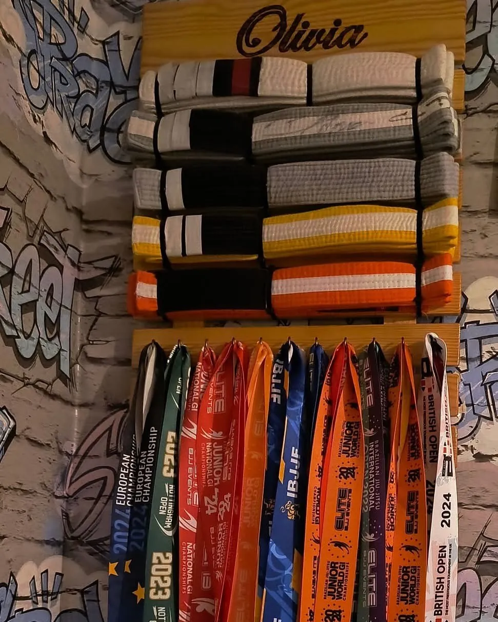 A wall rack carved with the name Olivia, holding her jiu-jitsu belts and medals from European, IBJJF, Junior World and British Open competitions