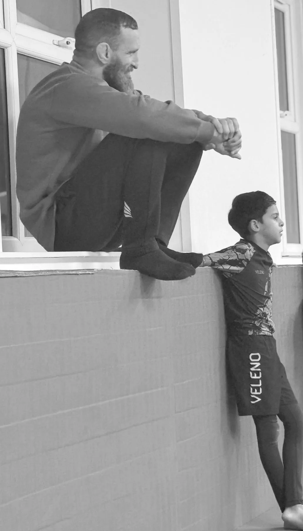A young boy in Dream Art training kit leaning against the wall watching the mat, with a coach sitting on the ledge beside him